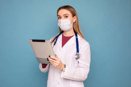 A female doctor in a protective mask writes down the diagnosis in a tablet on a blue backgroundの写真素材