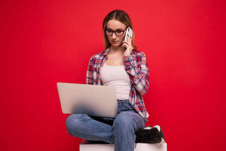 Cute young woman in stylish clothes talking on the phone and working with a laptop on a red backgroundの写真素材
