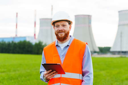 A male professional engineer smiles in a hard hat and a bright orange vest with a tablet against the...