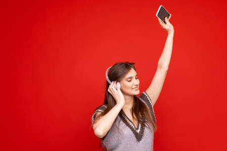 A young woman listens to music with headphones on a red background