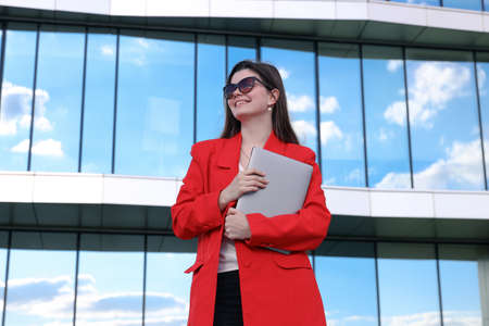 A businesswoman in a jacket poses with a laptop against the background of buildingsの写真素材