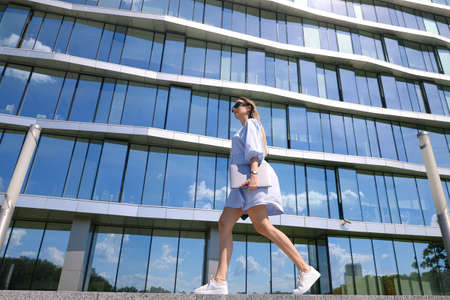A young woman walks with a laptop in the background of an office building