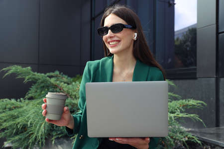 A young woman with a cup of coffee and a laptop poses on the street