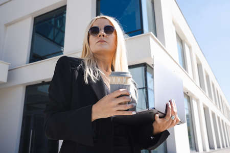 A young woman in a stylish suit with a laptop and a cup of coffee on the background of a business...