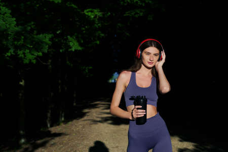 A sporty brunette with headphones and a bottle of water after jogging in the park. Tedious workouts outsideの写真素材