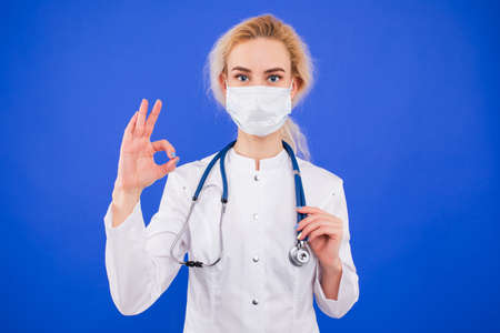 Portrait of a young female doctor in a protective mask shows the OK gesture on a blue backgroundの写真素材
