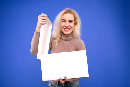 A surprised young woman opens a gift box on a blue background. The joy of a giftの写真素材