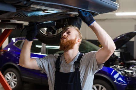 Inspection of the car suspension. A male engineer in uniform is standing under a car on a lift with...