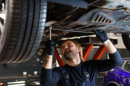 A male mechanic with a key unscrews the nuts on the bottom of the car that is on the lift....