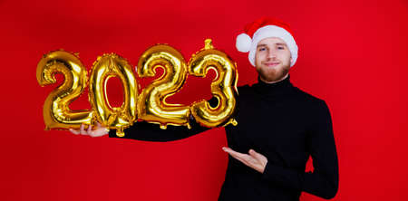 A man in a Santa Claus hat holds the shiny numbers of the new year 2023 on a red background. Family...