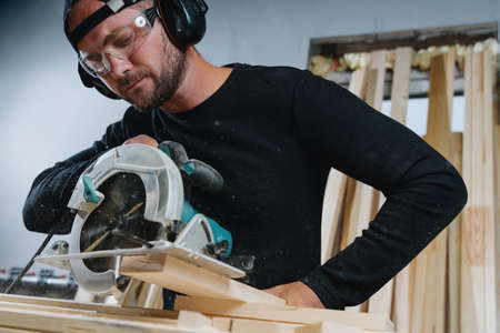Wood processing in the workshop. A bearded man in dark clothing and protective equipment cuts a...