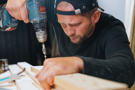 A carpenter man in dark clothes screws a self-tapping screw into a board with an electric screwdriver Professional furniture assembly in his workshopの写真素材