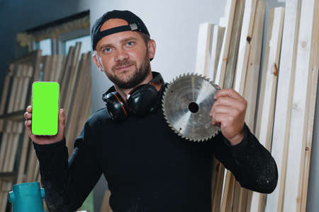 A carpenter man who is holding a phone with a green screen and a cutting disc from an electric saw. Buying Equipment Onlineの写真素材