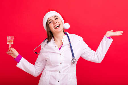 A nurse in a Santa Claus hat holds a glass of champagne happy New year on a red backgroundの写真素材