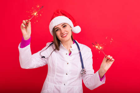 A joyful female doctor in a white coat with sparklers on a red background. Merry Christmas Greetingsの写真素材