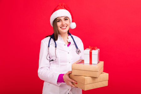 A female doctor in a Santa Claus hat holds boxes with gifts on a red background. Doctor's congratulations to patientsの写真素材