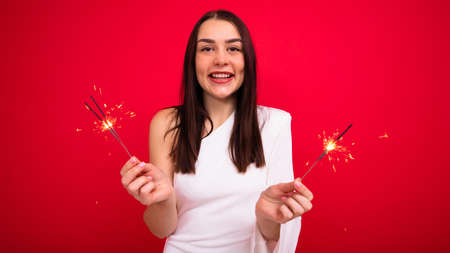 A cheerful brunette in an elegant white dress with sparklers on a red background in the studio. The...