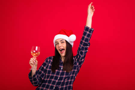 A smiling young woman in pajamas wearing a Santa Claus hat holds a glass of wine on a red background. New Year's Eve at homeの写真素材
