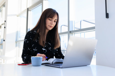 Working in a cafe - a brunette writes in a diary and looks at a laptop monitor.Remote work not in...