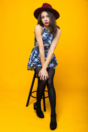 Fashion shooting of a young woman in a bright dress and hat on a yellow background. The model is sitting on a chair in the studio.の写真素材