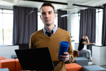 Portrait man with a laptop and a cup against the background of colleagues working in the officeの写真素材
