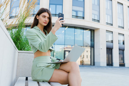 Portrait of a young woman with a laptop outside. Business woman while working on the street sitting on a benchの写真素材