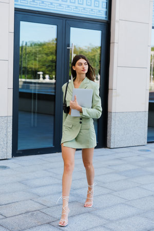 A young woman in a stylish skirt and jacket with a laptop in her hands goes to the office. street business portraitの写真素材
