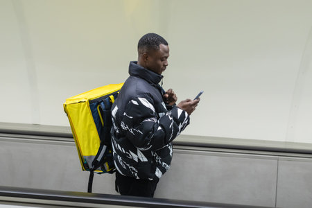 An African-American courier rides an escalator with a mobile phone in his hands. Yellow bright...