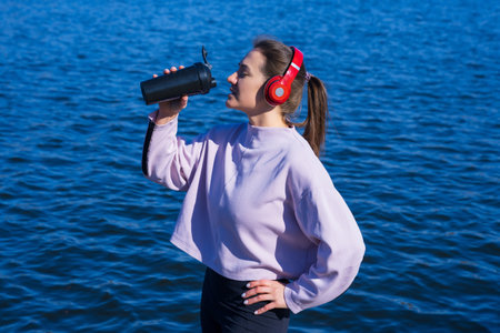 A young athletic woman drinks water after a workout outside.The athlete listens to music with headphonesの写真素材