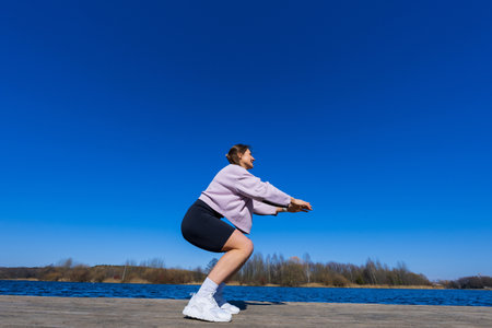 A young athletic woman squats during a workout. view of the pondの写真素材
