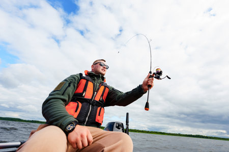 Porter of a fisherman sitting in a boat. A man with a spinning rod in his hands. Photo on a wide-angle lensの写真素材