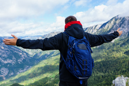 An unrecognizable man in a black jacket with a blue backpack stands with his hands raised to the side and enjoys the mountain landscapeの写真素材