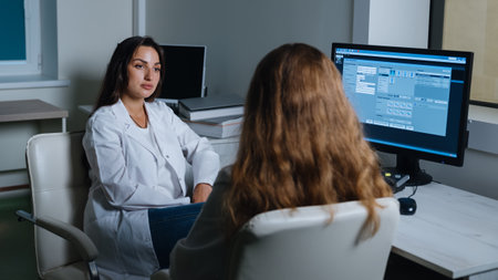 Two doctors are sitting at a table in front of a computer in an X-ray room and talking. Clinic with...