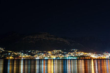 Night photography of a small town on the seashore against the backdrop of mountains. The lights of the city are reflected in the waterの写真素材