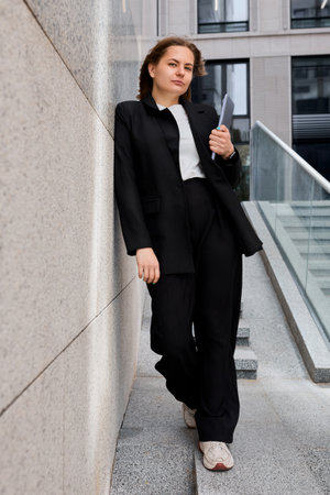 Professional businesswoman holds her laptop in her hands against the backdrop of a corporate office buildingの写真素材