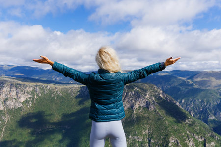 A young woman stands with her back to the camera and looks at the mountain landscape with her hands...