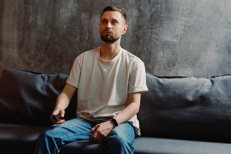 A handsome man with a beard in a white T-shirt and jeans holds a remote control from the TV and switches channelsの写真素材