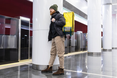 A stylishly dressed delivery man with a yellow backpack is waiting for a subway car. food delivery in a big cityの写真素材