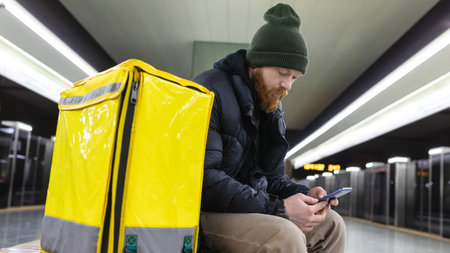 A courier with a yellow backpack holds a mobile phone and waits for a subway train on the platformの写真素材