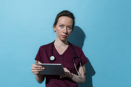 Young successful nurse woman dressed in burgundy uniform holding tablet.の写真素材