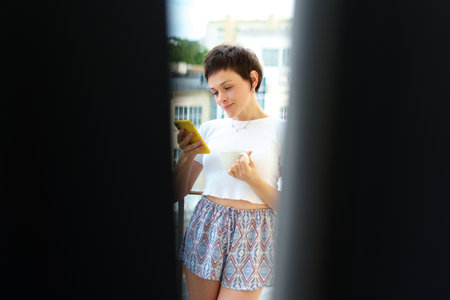 Morning at home. A young woman in pajamas is standing on the balcony, drinking coffee and listening to music against the background of the city.の写真素材