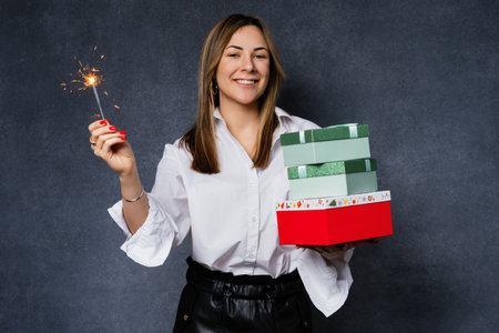 A happy young woman with gifts for the New Year and sparklers in the studio. The model looks at the camera on a dark backgroundの写真素材