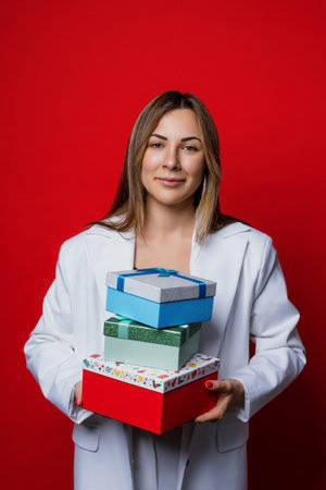 A woman in a stylish white jacket holds boxes of Christmas gifts on a red background. Winter holidays, vacationsの写真素材