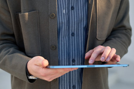 A man dressed in a shirt and jacket uses a tablet while standing on the street.の写真素材