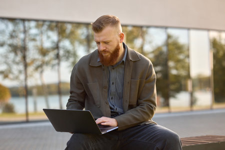 A European man with a red beard dressed in a shirt and jacket sits on a bench with a laptop.の写真素材