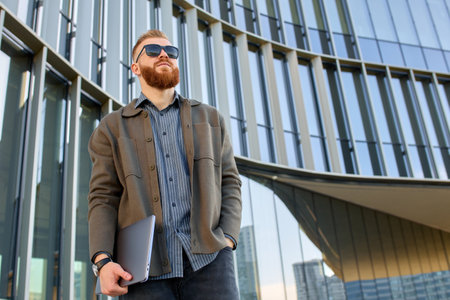 A man with a red beard dressed in a shirt and jacket stands at the entrance to a business center with a laptop.の写真素材