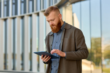 A man with a red beard dressed in a shirt and jacket checks his email on a tablet.の写真素材