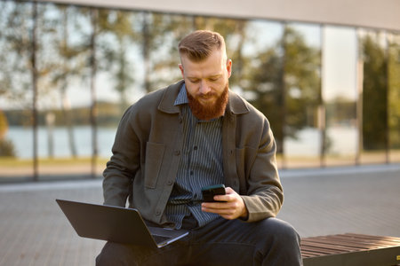 A 35-year-old man with a red beard sits on a bench with a laptop during lunch break.の写真素材