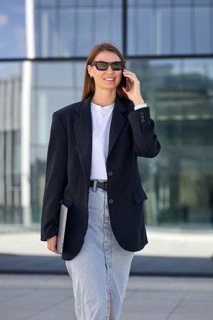 Stylish successful business woman dressed in a skirt and jacket speaks on the phone with business partners while walking down the street.の写真素材
