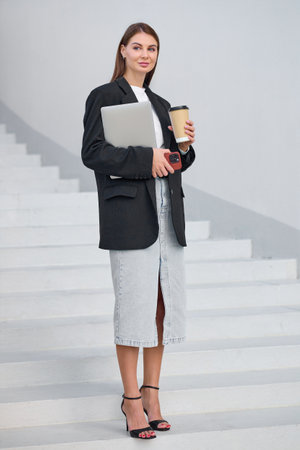 Young woman with brown hair looks successful and confident holding a laptop in her hands while standing on the street.の写真素材